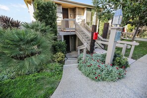 Un edificio residencial con una escalera que conduce a un porche, rodeado de abundante vegetación y un cantero de flores.