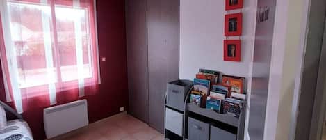 Een kamer met een bed, een radiator, een raam met gordijnen, een kledingkast en een klein boekenrek met boeken.