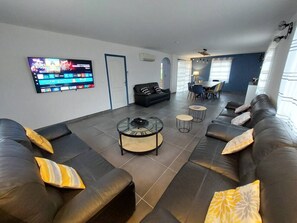 A modern living room with a large flat-screen TV mounted on the wall, a black leather sofa with yellow and white cushions, a glass coffee table, and a dining area with a table and chairs.