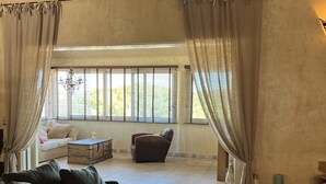 A spacious living area with a sofa, a small table, and a chair. There is a window with curtains, and a chandelier is visible. - Villa Ghjulia (Porto-Vecchio)