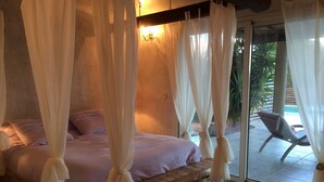 A bedroom with a stone bench, a bed with white curtains, and a view of a pool and lounge chair outside. - Villa Ghjulia (Porto-Vecchio)
