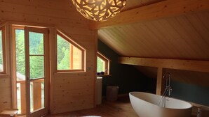 Puinen mökki, jonka sisätiloissa on suuri sänky, kylpyamme ja ikkuna, josta avautuu näkymä metsään.