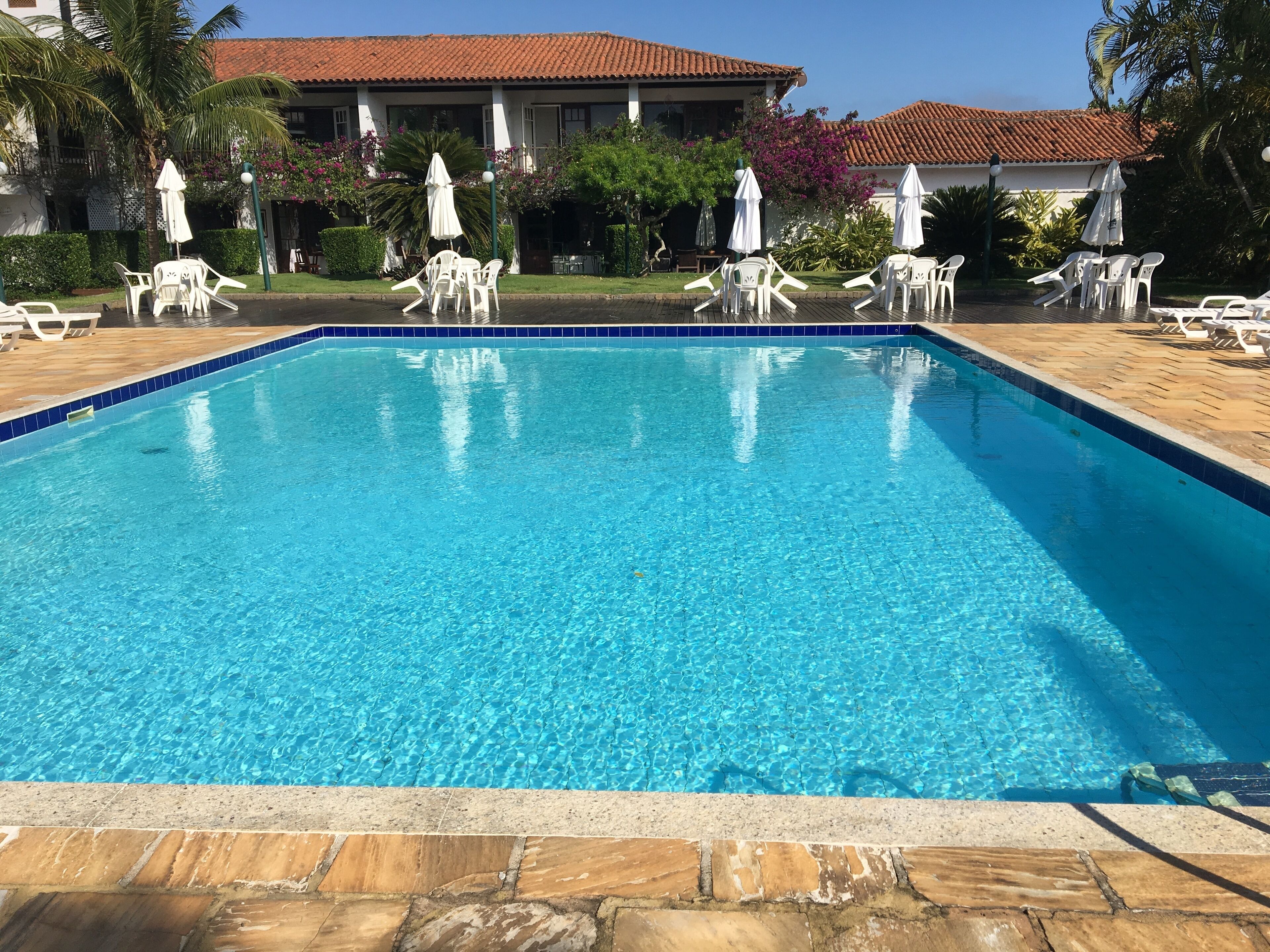 A clear blue swimming pool with white lounge chairs and umbrellas, surrounded by a stone deck and a building with a tiled roof.