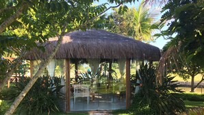 A thatched-roof structure with glass walls, surrounded by lush greenery and a paved path.