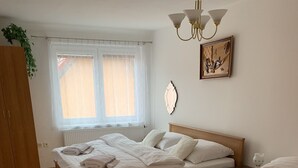 A bedroom with two beds, a wooden wardrobe, a radiator, a window with sheer curtains, a wall-mounted light fixture, and a framed picture.