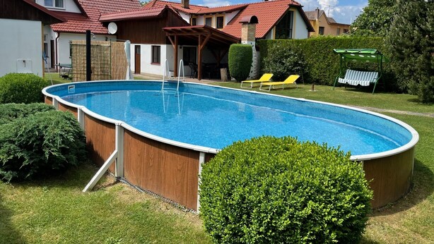 A residential swimming pool with a wooden deck, surrounded by well-maintained lawns and hedges.