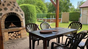 An outdoor patio with a stone-built barbecue grill, a table with chairs, and a neatly trimmed hedge in the background.