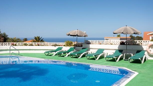 A pool area with green lounge chairs and umbrellas, a clear sky, and a view of the sea. - Beautiful Apartment in Parque las Flores (Puerto de la Cruz)