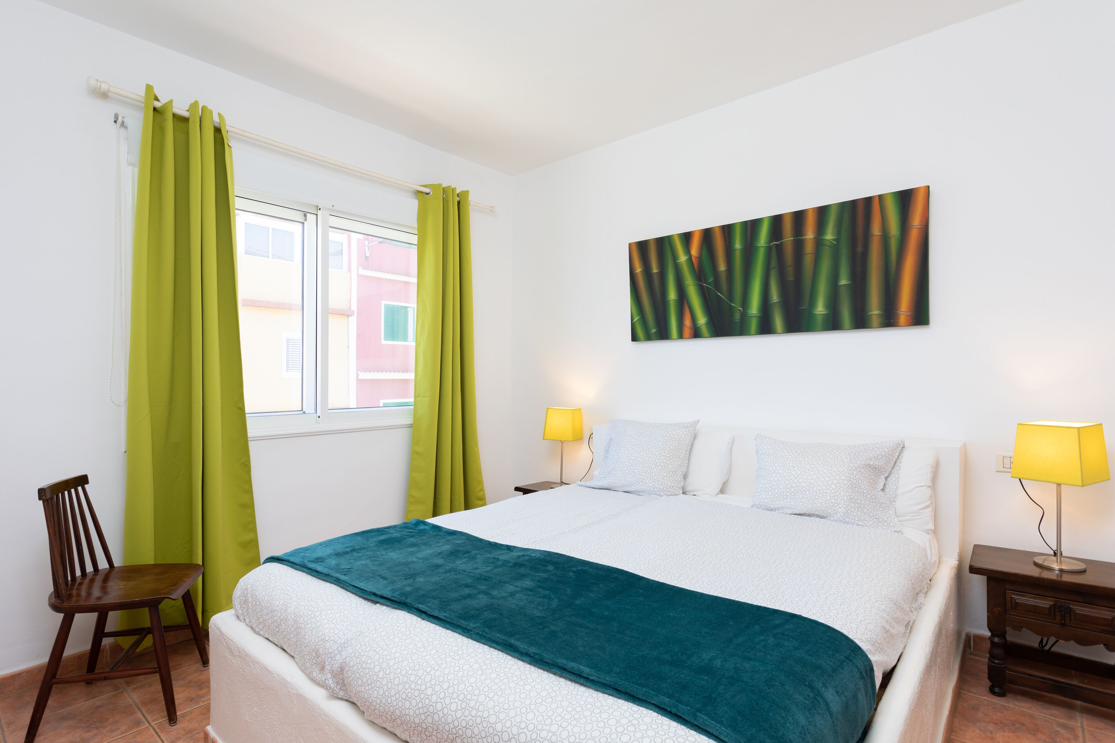 A bedroom with a bed, green curtains, a wooden chair, a nightstand with a lamp, and a wall art piece.