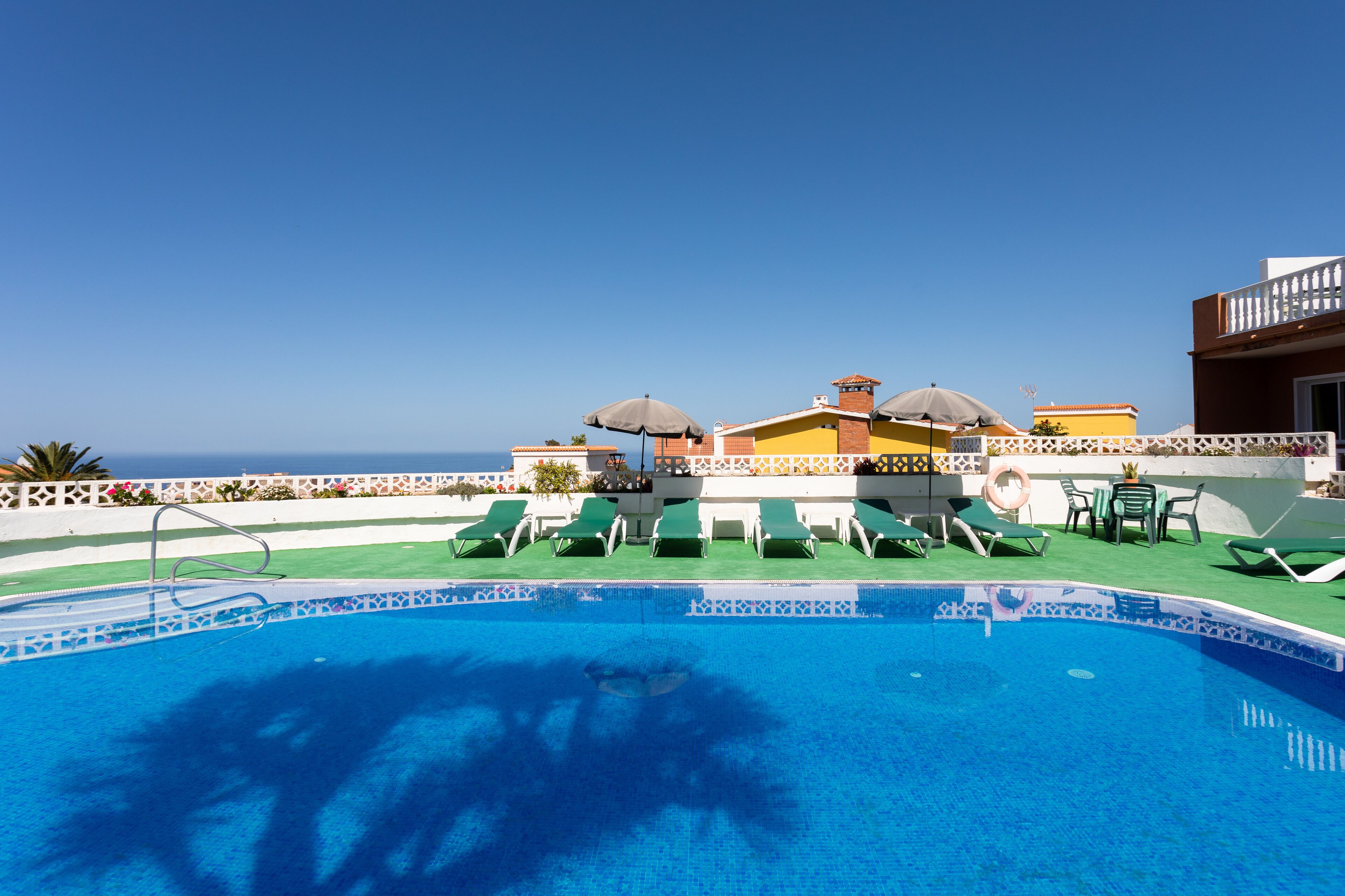 A pool area with lounge chairs and umbrellas, a clear view of the ocean, and a building with a white fence and colorful tiles.