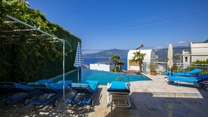 Un espace piscine doté de chaises longues bleues et de parasols, offrant une vue panoramique sur la mer et les montagnes.