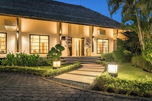 A well-lit building with a thatched roof, a paved pathway leading to the entrance, and lush greenery surrounding the property. - Luxury Beach Villa with Infinity Pool. (Singaraja)