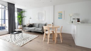 Ein modernes Wohnzimmer mit einem grauen Sofa, einem runden Holztisch mit Stühlen, einem weißen Couchtisch, einem großen Fenster mit grauen Vorhängen, einer Topfpflanze und einer weißen Wand mit einem gerahmten Bild und einer Vase mit Blumen.