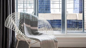 Ein weißer, filigraner Stuhl mit gemustertem Überwurf, platziert bei einem Fenster mit Blick auf eine blaue Backsteinmauer.
