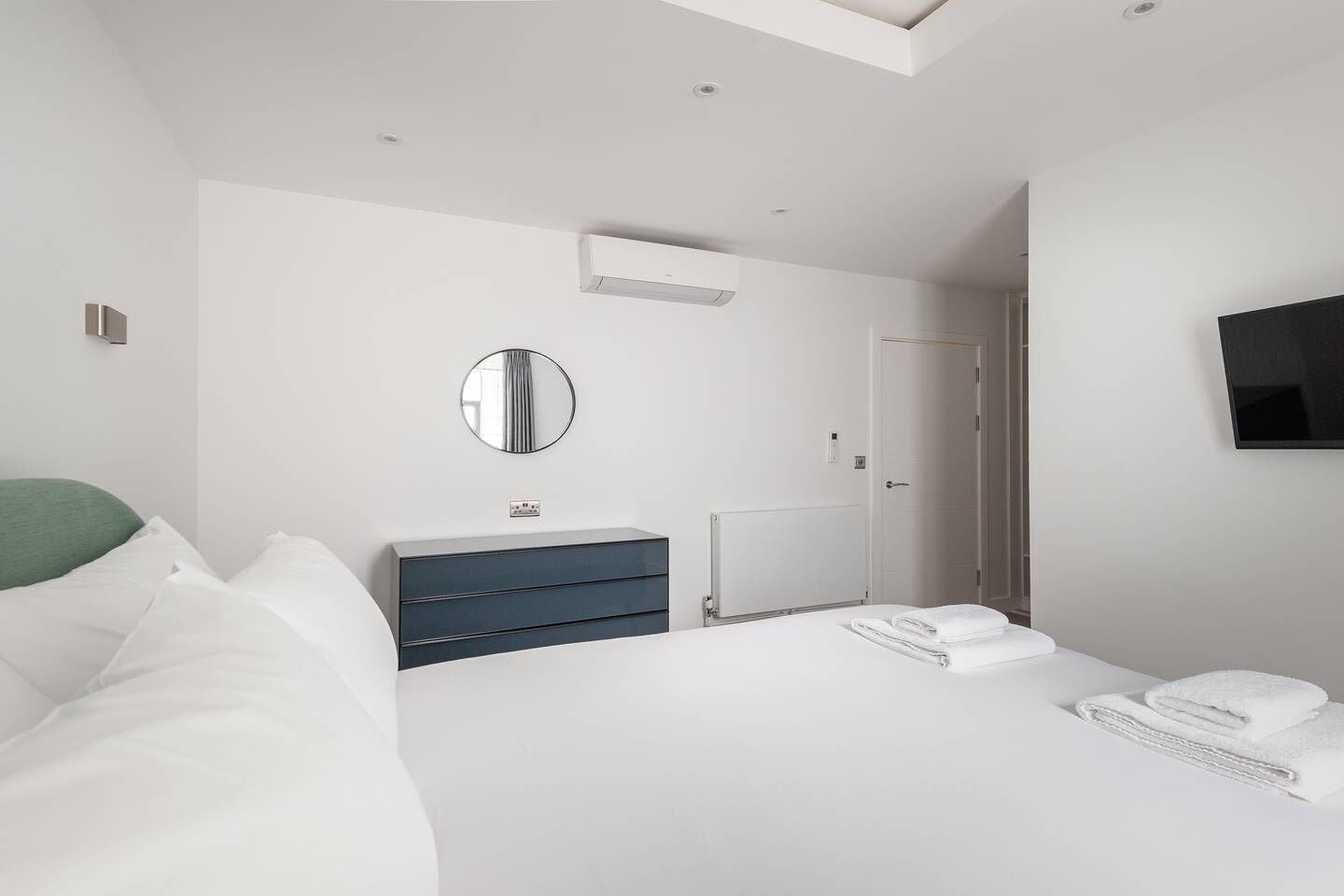 A modern hotel room with a large bed, a bedside table, a mirror, a wardrobe, a TV, and a door leading to a bathroom.