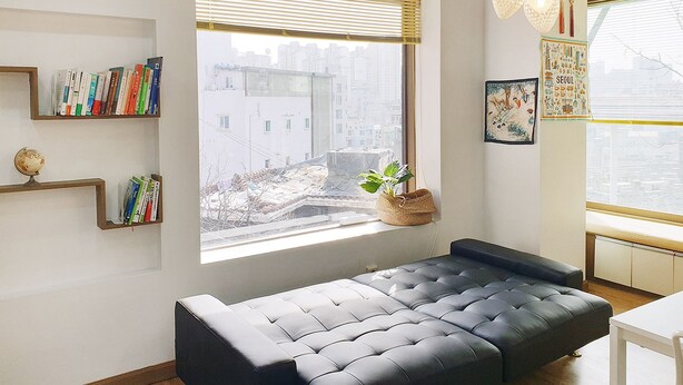 A modern living room with a black sofa, a bookshelf, a window with blinds, and a hanging light fixture. - Cheese Guesthouse - Hostel (Seoul)