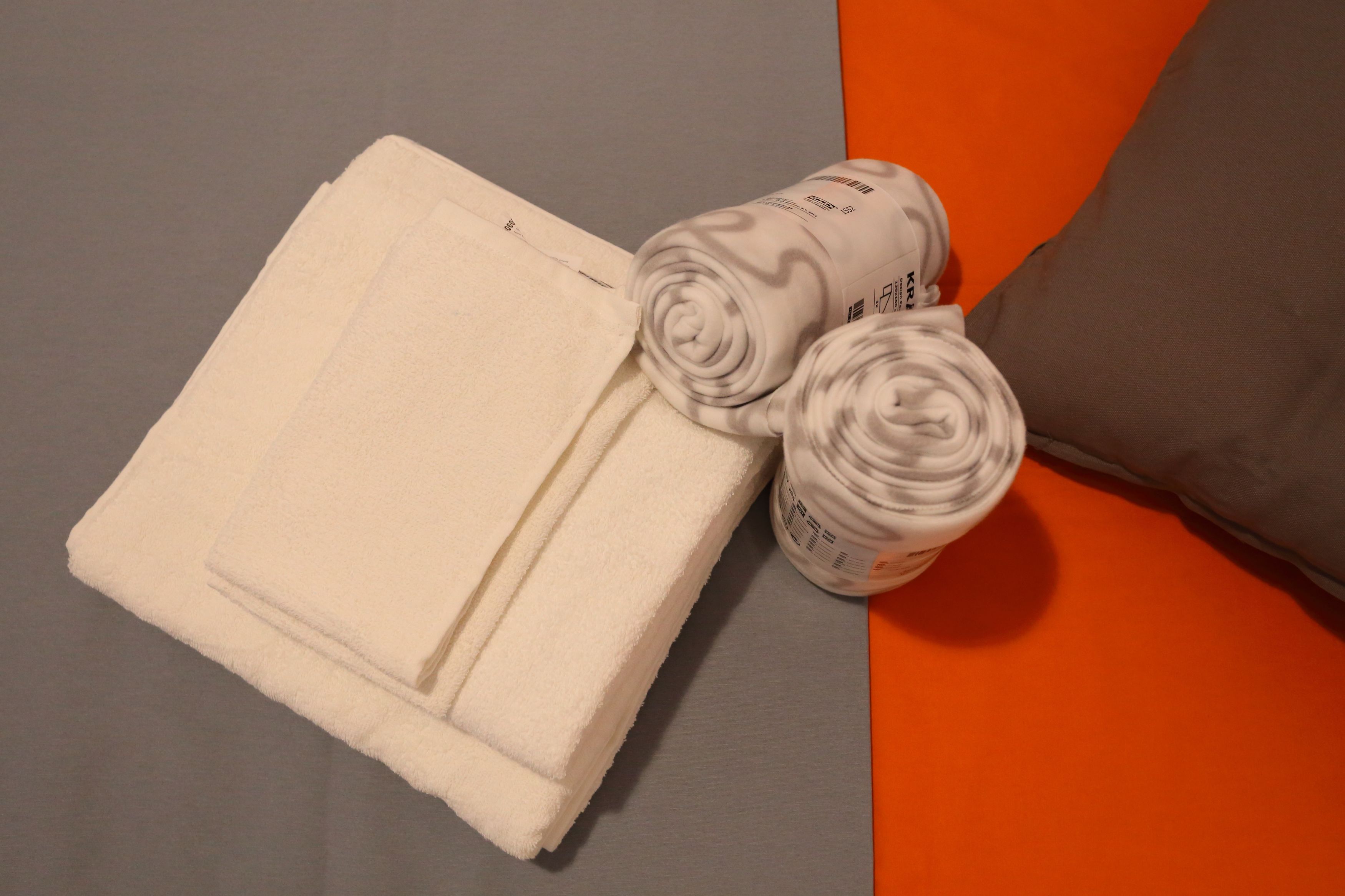 Una toalla blanca doblada con cuidado y dos toallas enrolladas sobre una cama con un cojín gris y naranja.