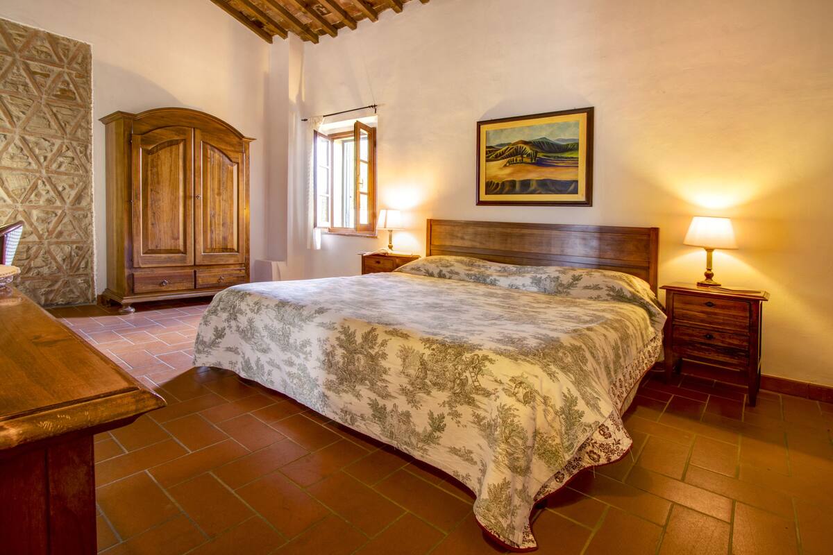 Une chambre avec un grand lit, une armoire en bois, deux tables de chevet avec des lampes, une fenêtre donnant sur un beau paysage et un tableau accroché au mur.