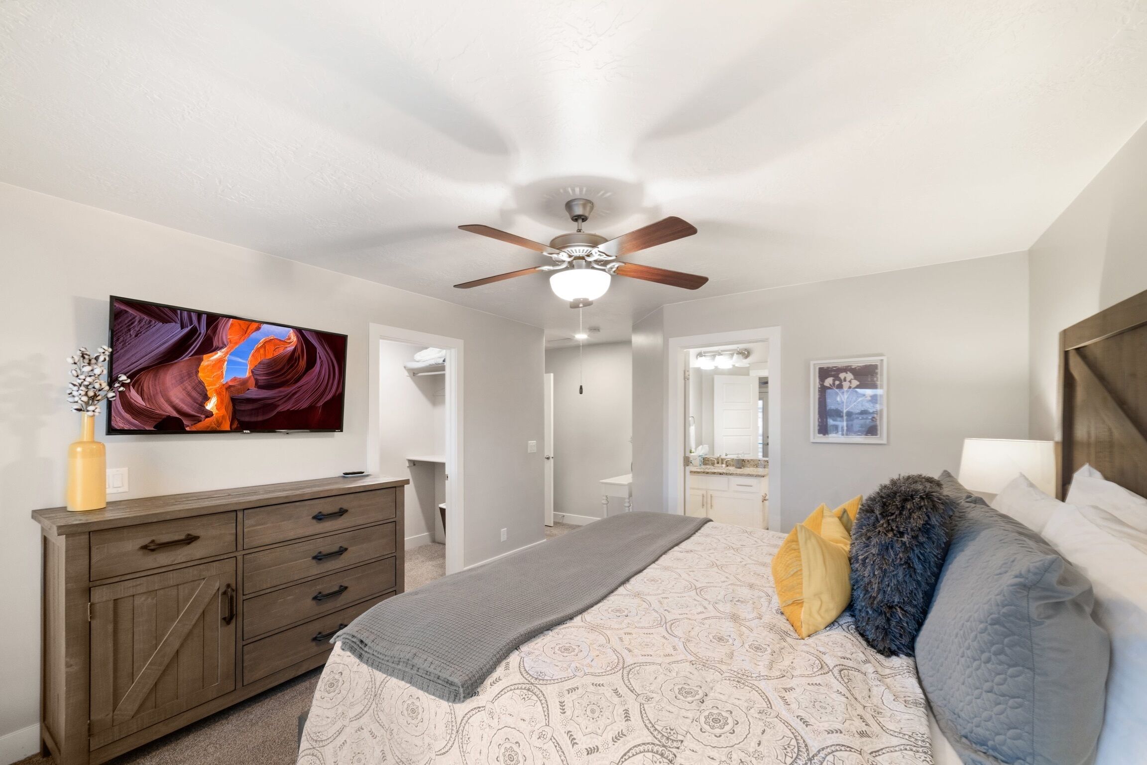 Une chambre à coucher avec un lit, un ventilateur de plafond, un téléviseur fixé au mur, une commode et une salle de bain visible à travers une porte ouverte.