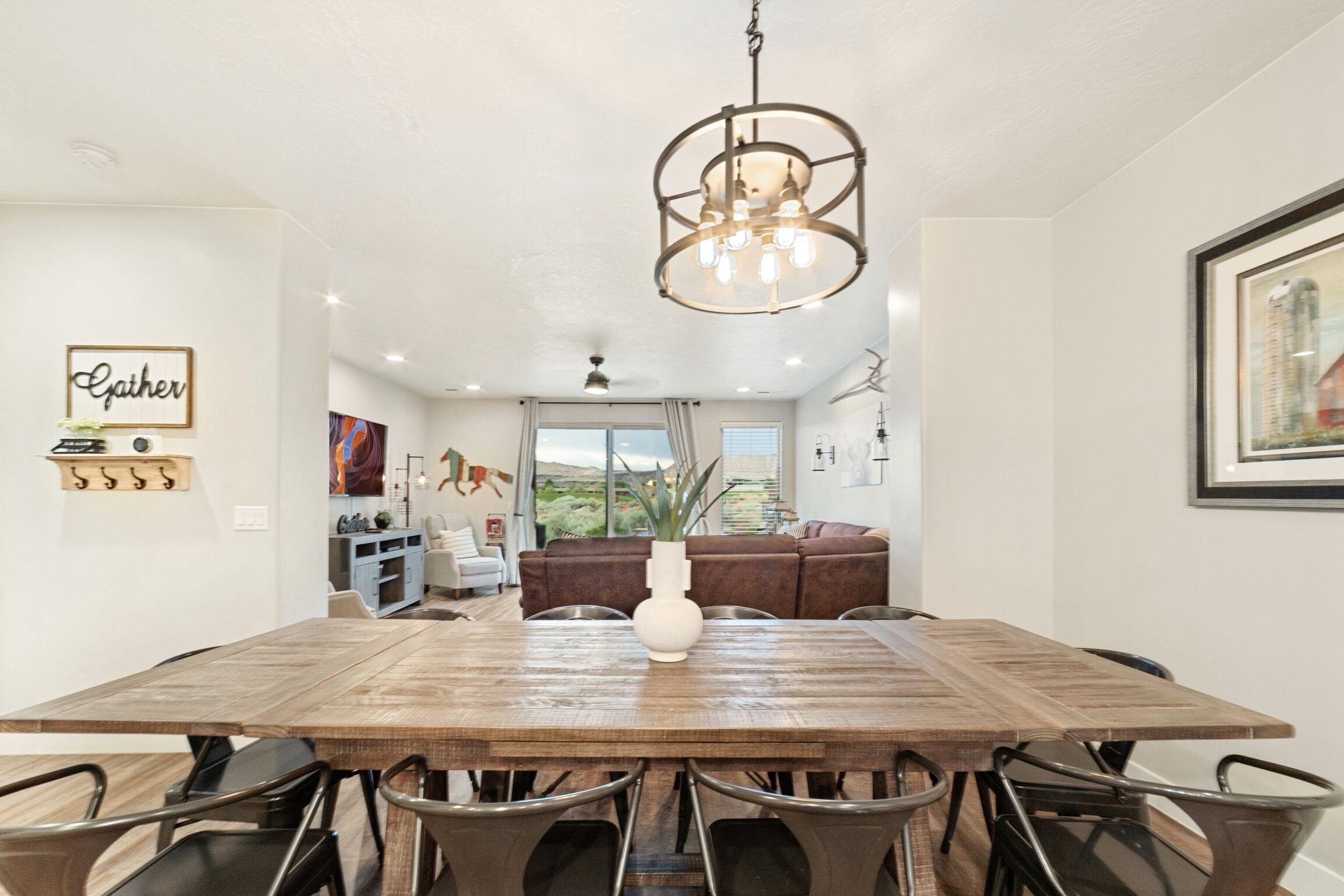A dining area with a wooden table, metal chairs, a hanging light fixture, and a view of a living room with a brown sofa and a vase.