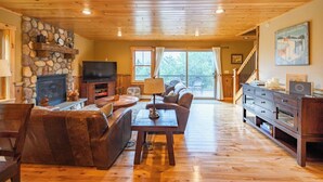 Ein gemütliches Wohnzimmer mit Kamin aus Stein, Holzmöbeln, einem Fernseher und einer Treppe, die zu einem Balkon führt.