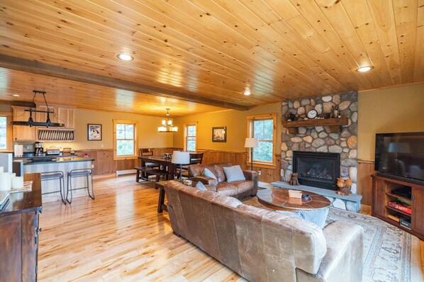 A spacious living area with a wooden ceiling, a stone fireplace, a large TV, and a cozy seating arrangement.