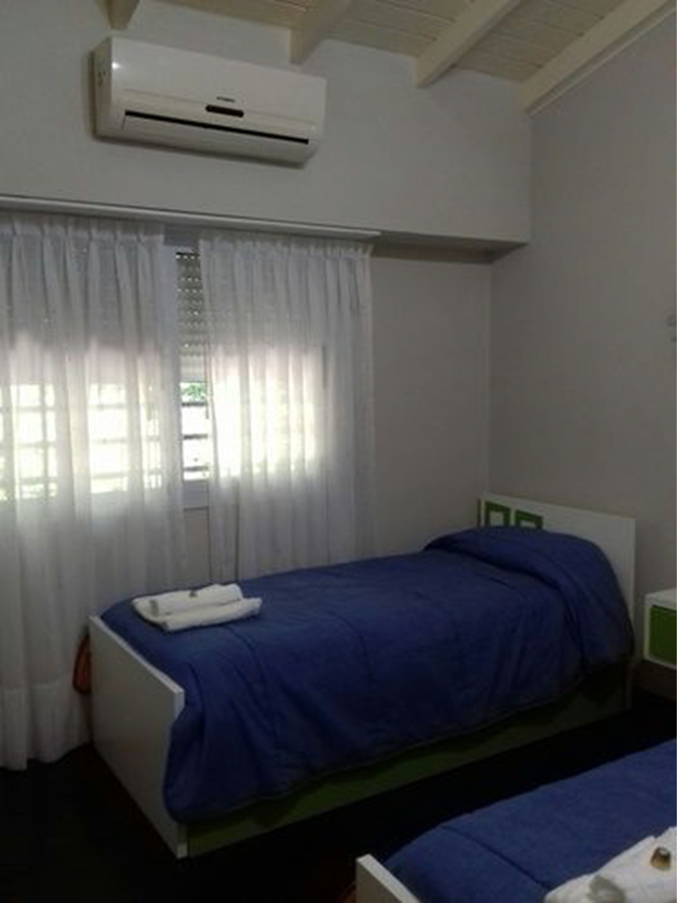 A single-bed room with a blue bedspread, white curtains, and an air conditioning unit on the wall.