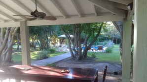Um pátio externo com mesa e cadeiras de madeira, um ventilador de teto e vista para uma piscina e área de jardim.