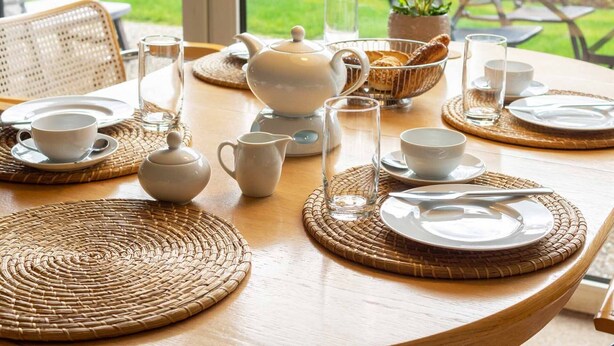 A breakfast setting with a table set for two, including plates, cups, a teapot, and a basket of pastries.