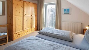 A bedroom with a large wooden wardrobe, a bed with two pillows, a window with a view of buildings, and a radiator.