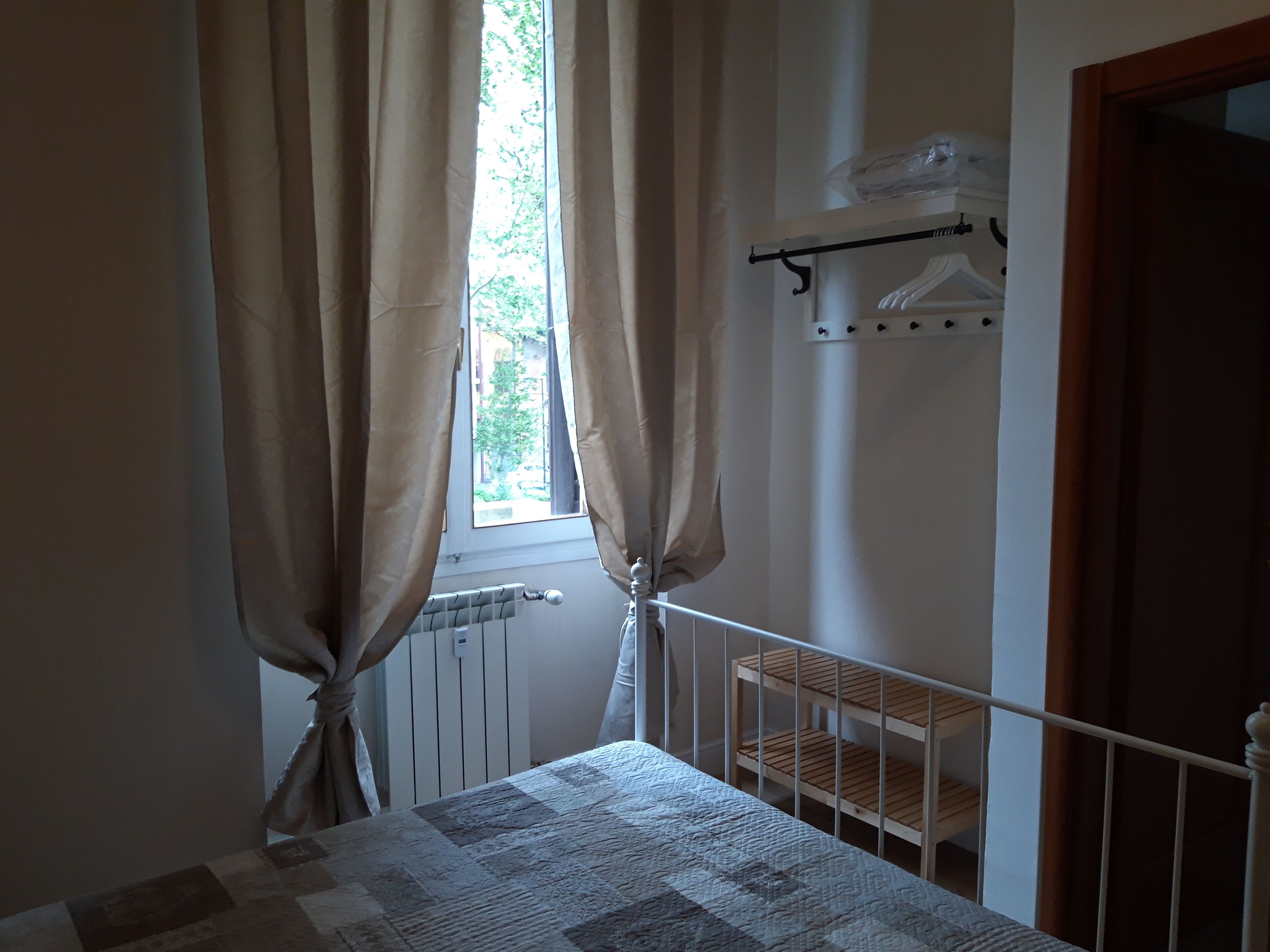 A bedroom with a bed, a radiator, a window with curtains, a wooden shelf, and a coat rack.