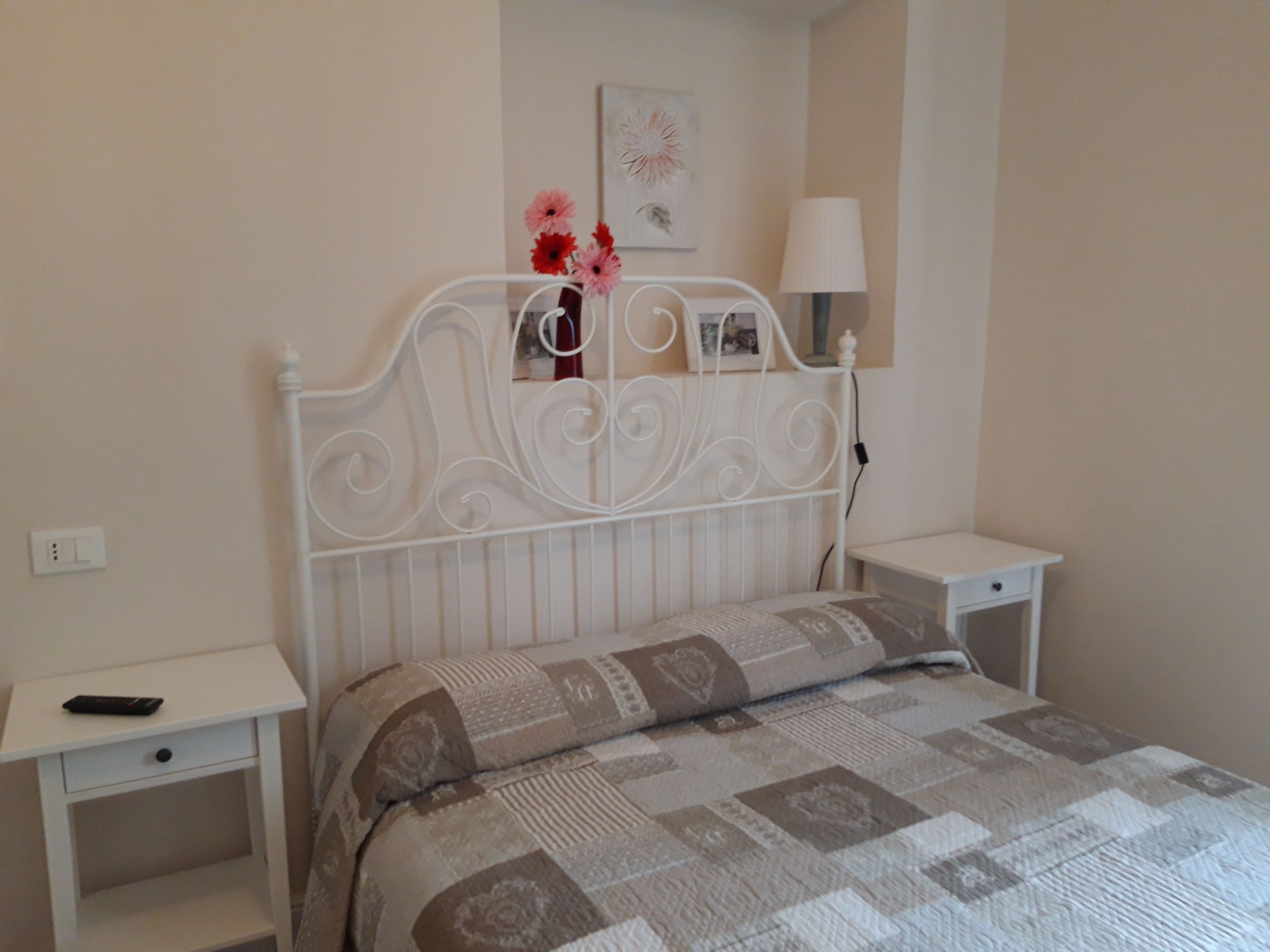 A bedroom with a white metal headboard, a patterned bedspread, a bedside table with a lamp, and a small shelf with flowers.