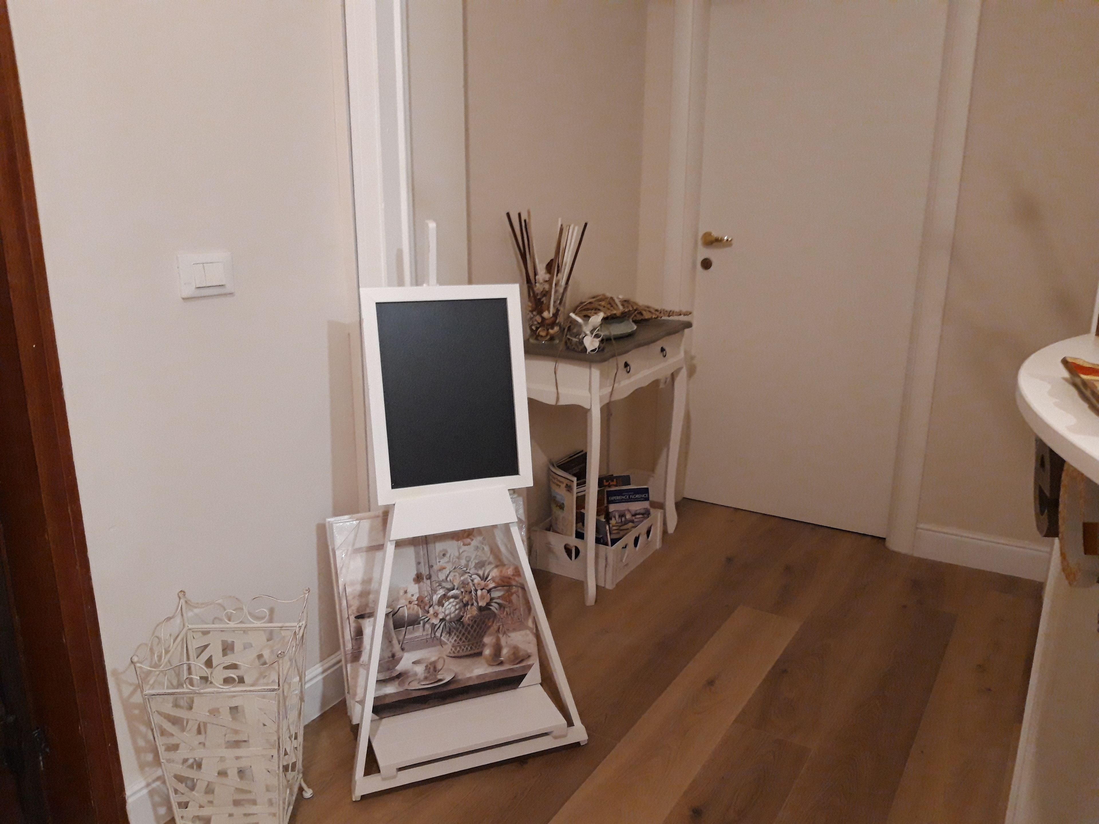 Una stanza con un cavalletto bianco su cui è tesa una tela vuota, un tavolino con oggetti decorativi e un armadietto bianco pieno di libri.