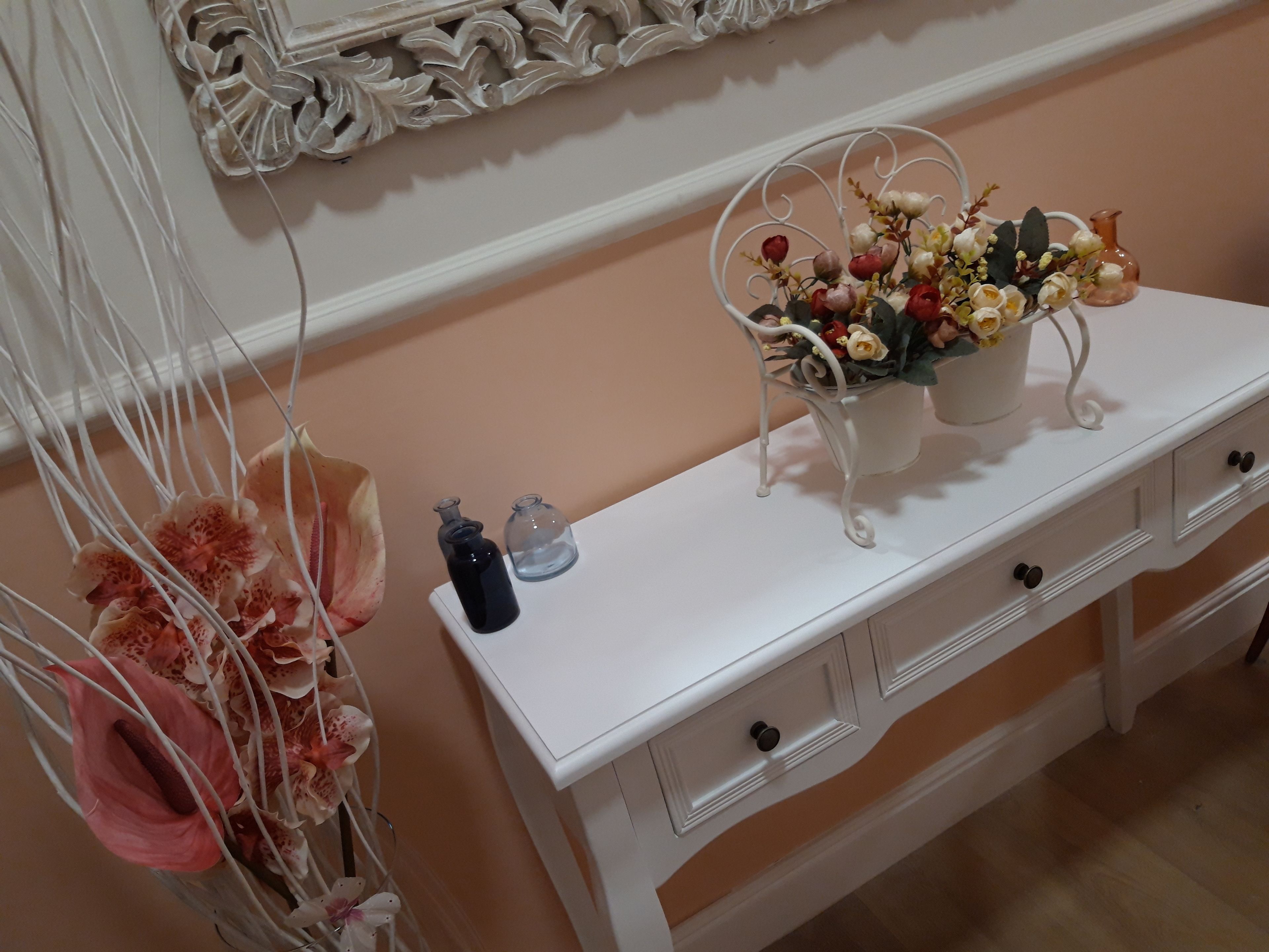 Ein weißer Schminktisch mit Schubladen, ein Spiegel darüber und eine Vase mit Blumen darauf.