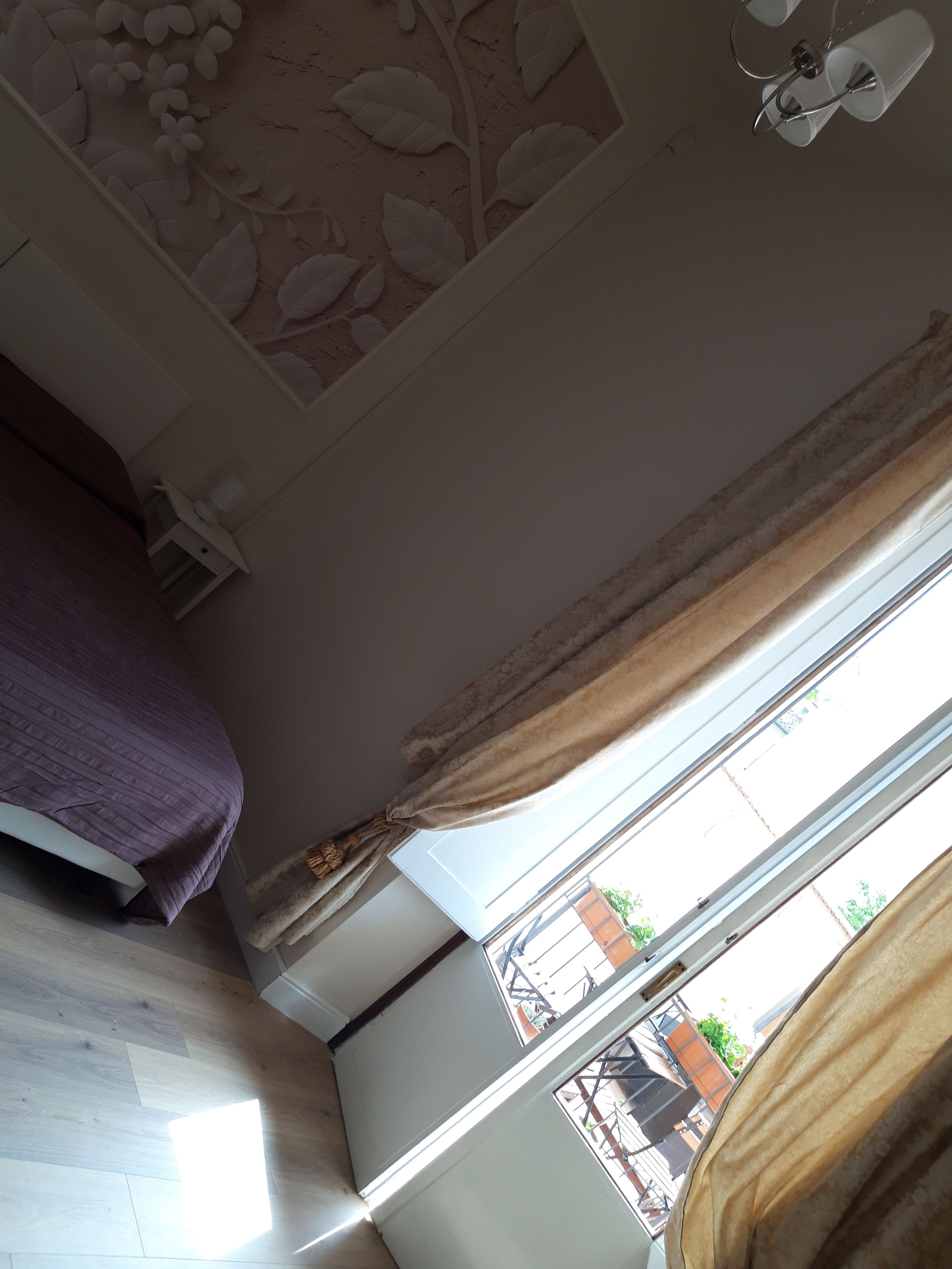 Un dormitorio con cama, techo decorado con motivos florales, ventana con cortinas y vistas al exterior.