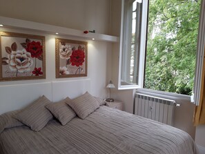 A bedroom with a bed, two wall-mounted floral paintings, a bedside lamp, and a window with a view of greenery. - Il Sorriso (Florence)