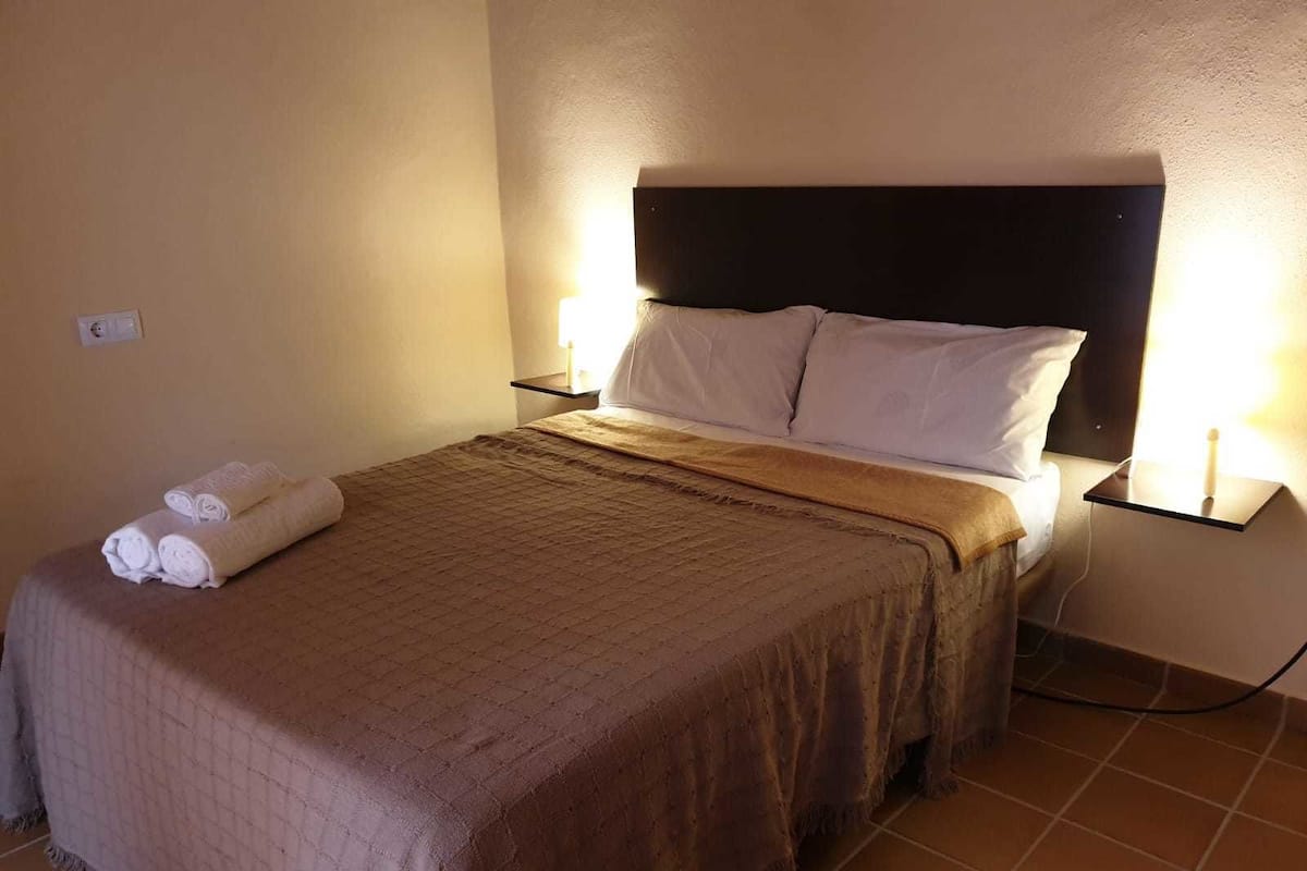 A neatly made bed with a brown blanket and pillows, two bedside tables with lamps, and a tiled floor.