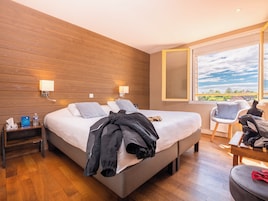 Quarto de hotel com uma cama grande, paredes de painel de madeira, mesa de cabeceira com luminária, uma cadeira e vista para o exterior.