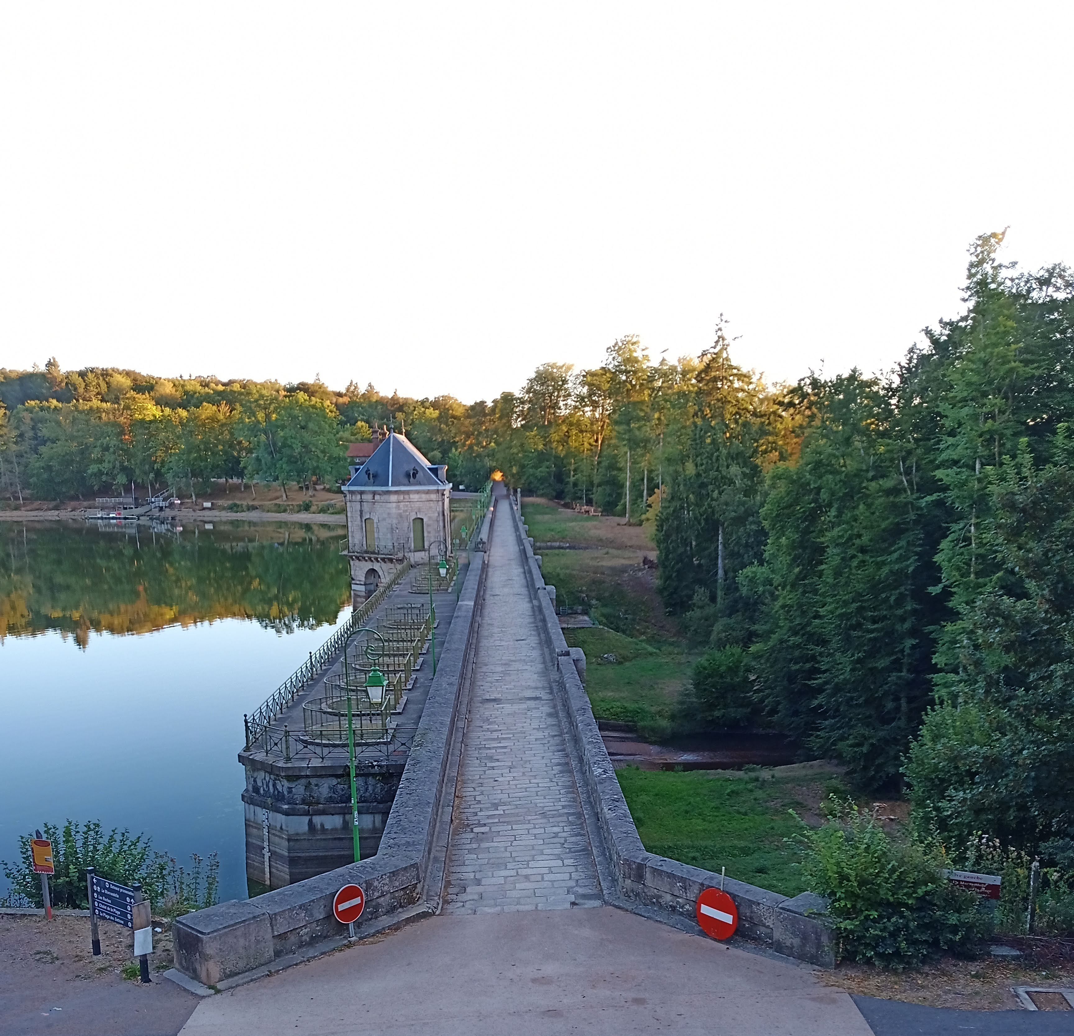A long bridge leading to a small building with a pointed roof, surrounded by trees and a calm body of water.