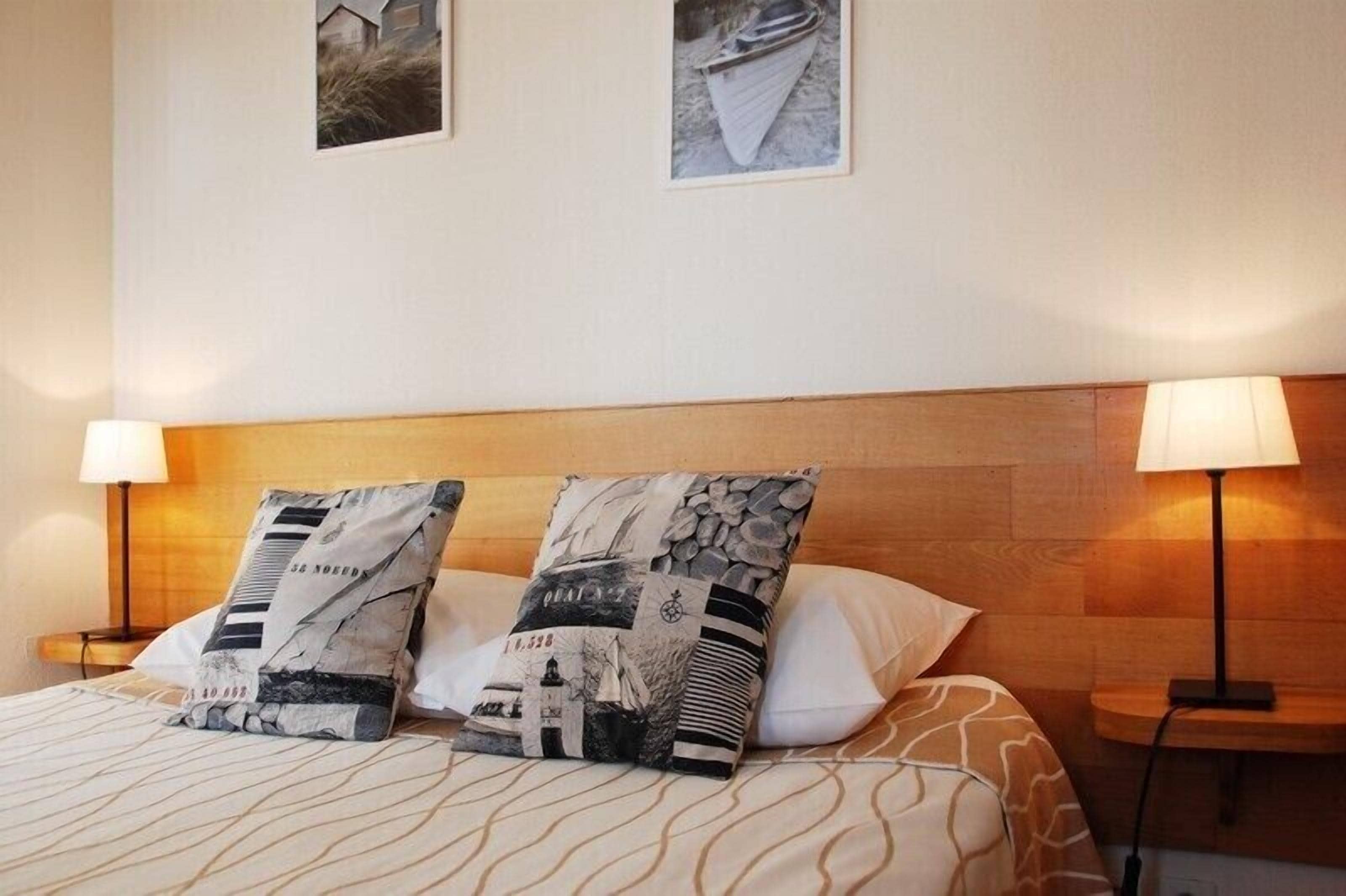 A neatly made bed with patterned pillows, two wall-mounted lamps, and two framed pictures above the bed.
