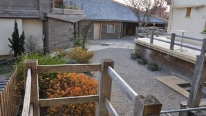 A courtyard with a wooden fence, a small garden with orange foliage, and a building with a grey roof and wooden siding. - Le Relais des Lacs (Planchez)