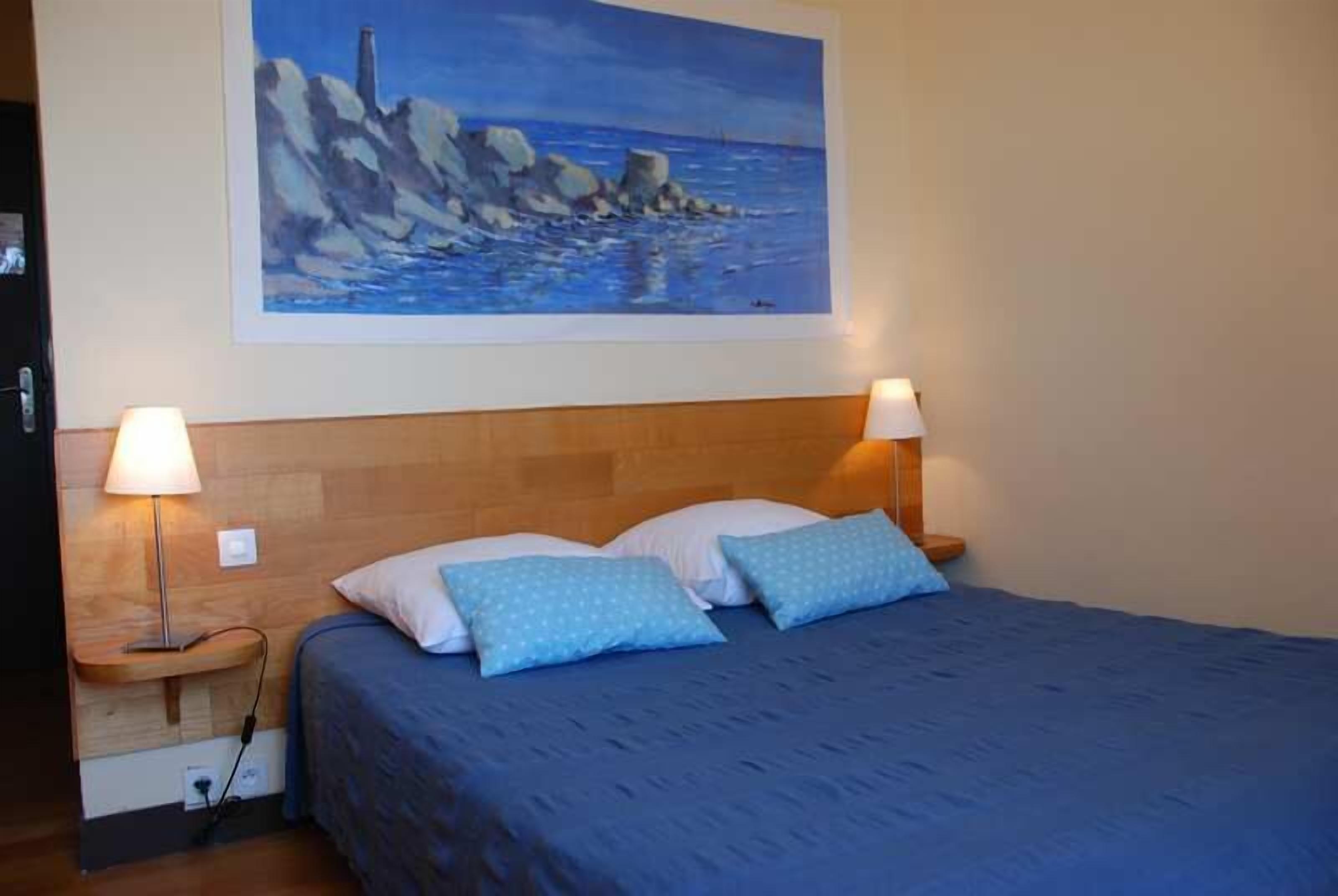 Un dormitorio con cabecera de madera, un cuadro de una escena costera, dos lámparas de noche y una colcha azul.
