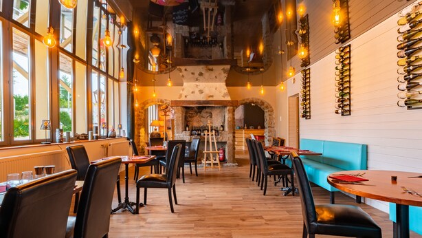 A restaurant interior with wooden floors, black leather chairs, and tables. There are hanging light bulbs and a wine rack on the wall. - Le Relais des Lacs (Planchez)