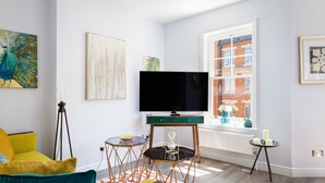 Un salon moderne avec un téléviseur à écran plat, un canapé jaune et turquoise, et une grande fenêtre donnant sur la ville.