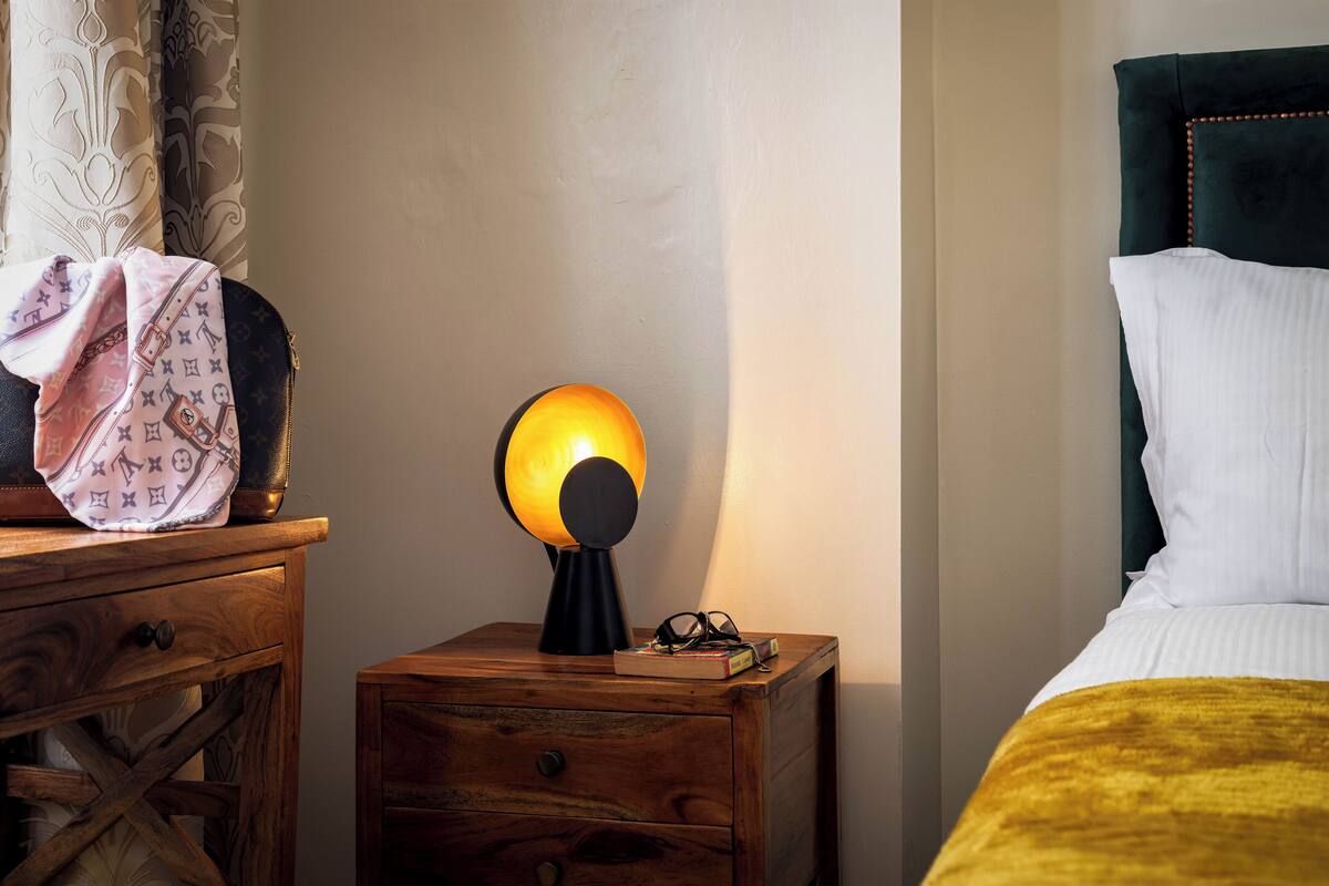 Une table de chevet en bois, avec une lampe, une paire de lunettes et un sac rose à motifs.