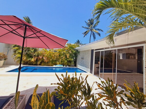 A swimming pool with a red umbrella, surrounded by tropical plants and palm trees.