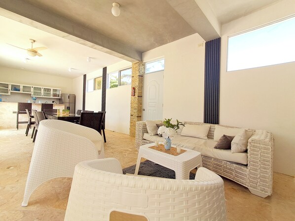 A spacious living area with a white sofa, a coffee table, and wicker chairs. There is a kitchen in the background with cabinets and a refrigerator.