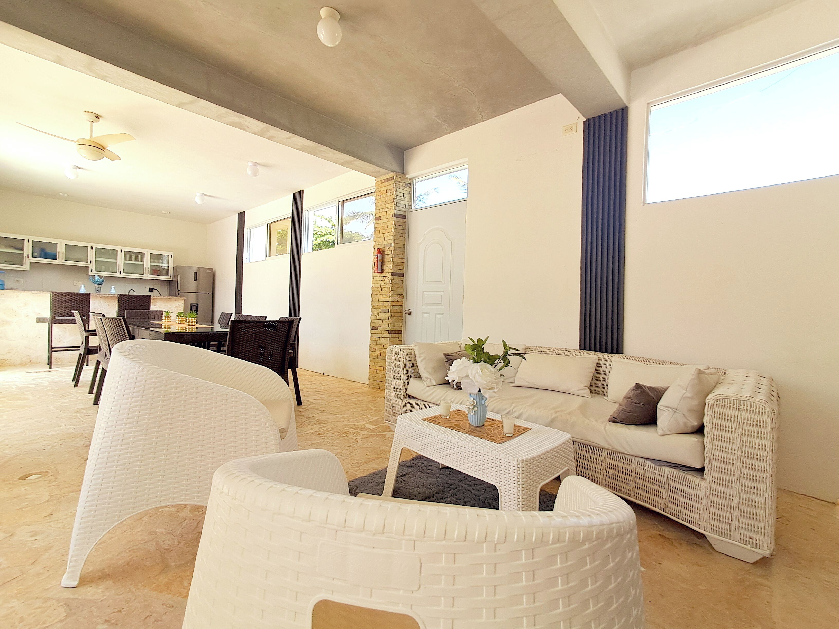 A spacious living area with a white sofa, a coffee table, and wicker chairs. There is a kitchen in the background with cabinets and a refrigerator.