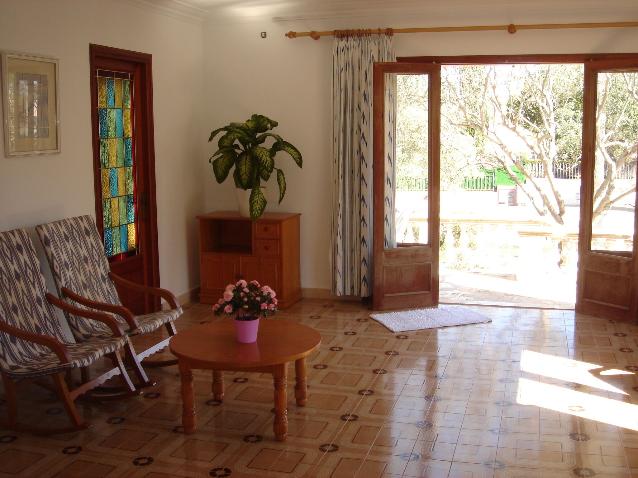 A room with a patterned floor, a wooden table with a vase of flowers, a rocking chair, a cabinet, and a door leading outside.