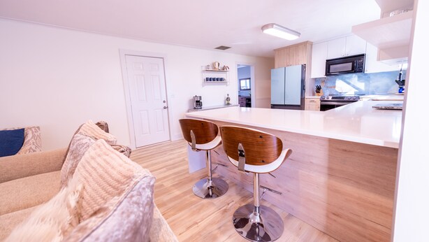 A modern kitchen with a central island, two bar stools, a wooden floor, and a white door. - .6 Walk to Carmel Midtown. Quiet and secure neighborhood. Plus massage chair! (Carmel)